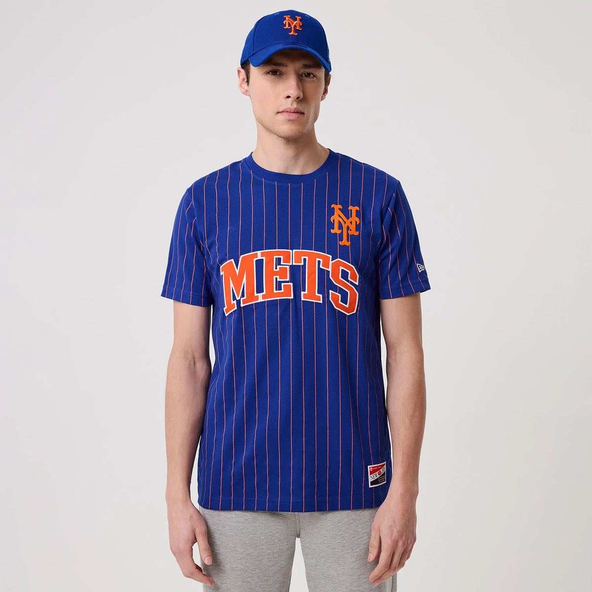 New Era New York Mets MLB Pinstripe Blue T-Shirt, front view on-model showing the orange METS lettering across the chest, a small orange embroidered Mets logo on the left chest, and fine vertical pinstripes on the blue fabric.