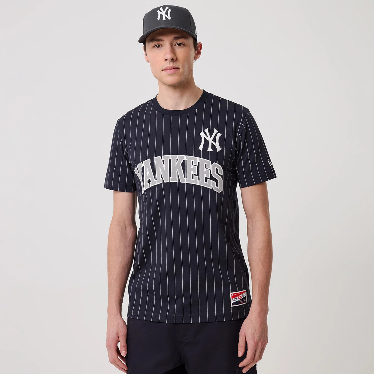 New Era New York Yankees MLB Pinstripe Navy T-Shirt, front view on a model with a grey cap, featuring white pinstripes on a navy background and an embroidered Yankees logo on the chest above Yankees lettering.