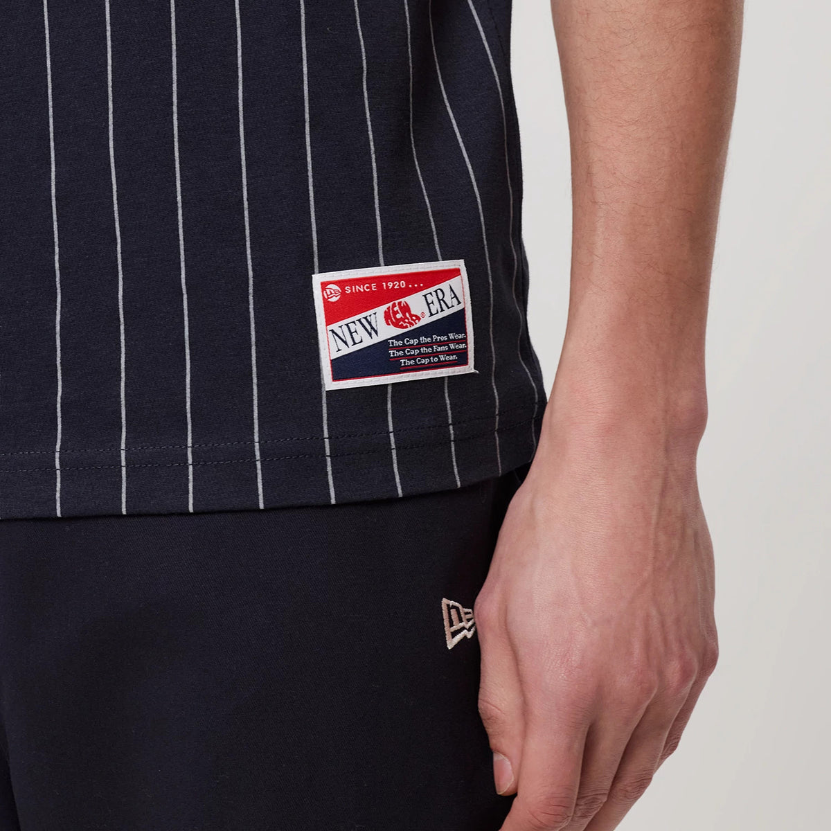 New Era New York Yankees MLB Pinstripe Navy T-Shirt, front view showing thin white vertical pinstripes and a square patch near the hem with New Era branding, worn by a model with dark trousers featuring an embroidered New Era logo.