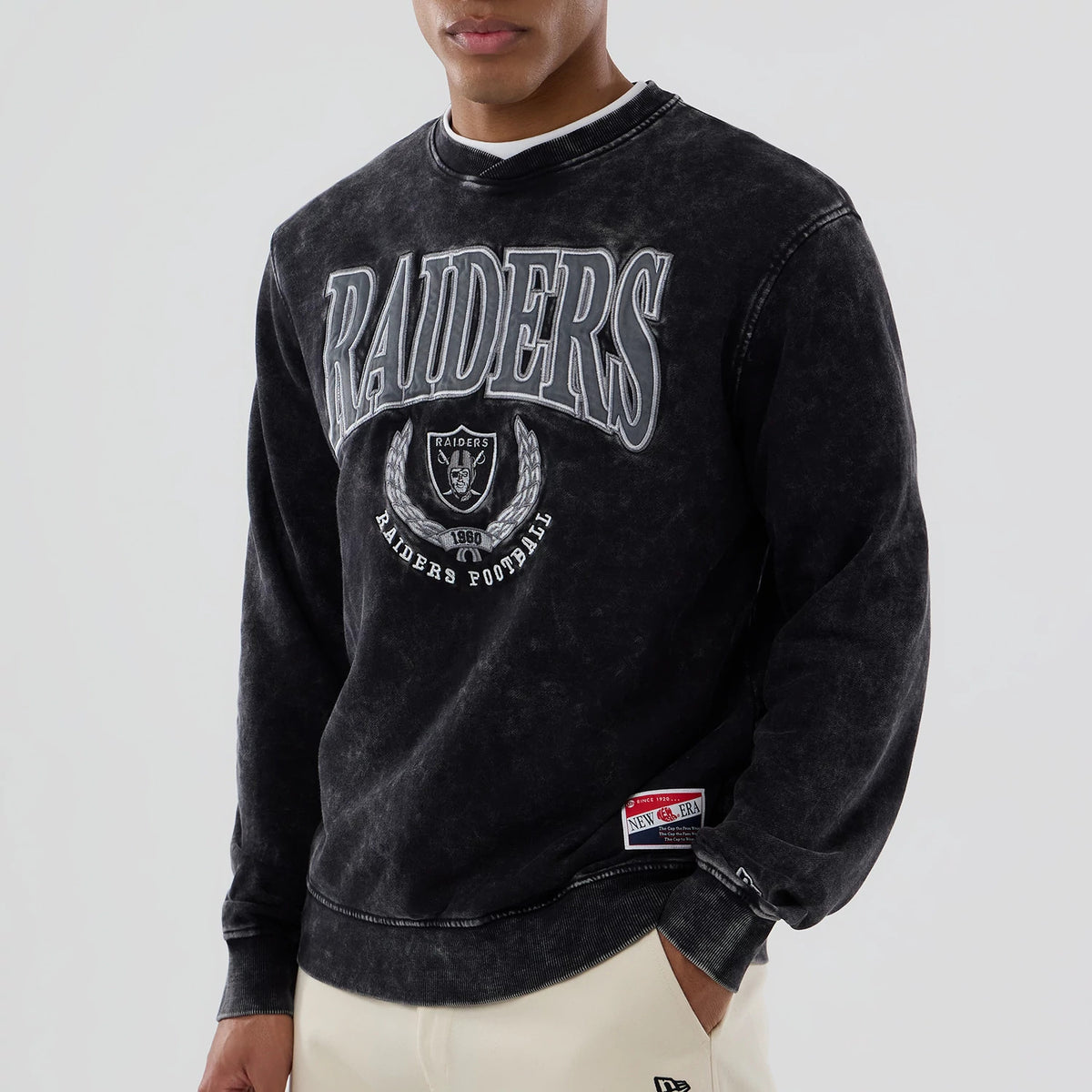 New Era Las Vegas Raiders NFL Laurel Wreath Black Crew Neck Sweater, front view on a model featuring large raised silver Raiders lettering and logo on the chest, with a New Era patch on the lower left hem.