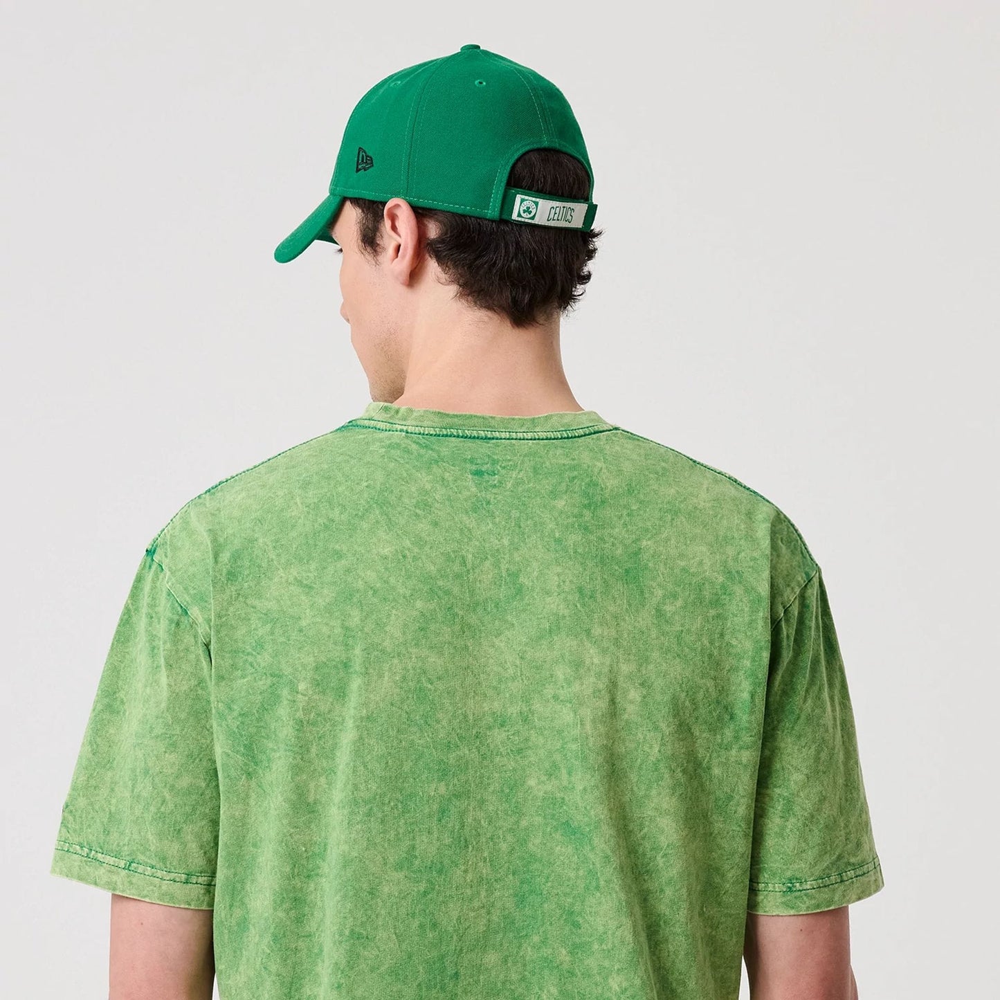 New Era Boston Celtics NBA Green T-Shirt, back view on a model wearing a matching green cap, showing a solid green back with stitching details and a New Era logo embroidered on the cap's side panel.