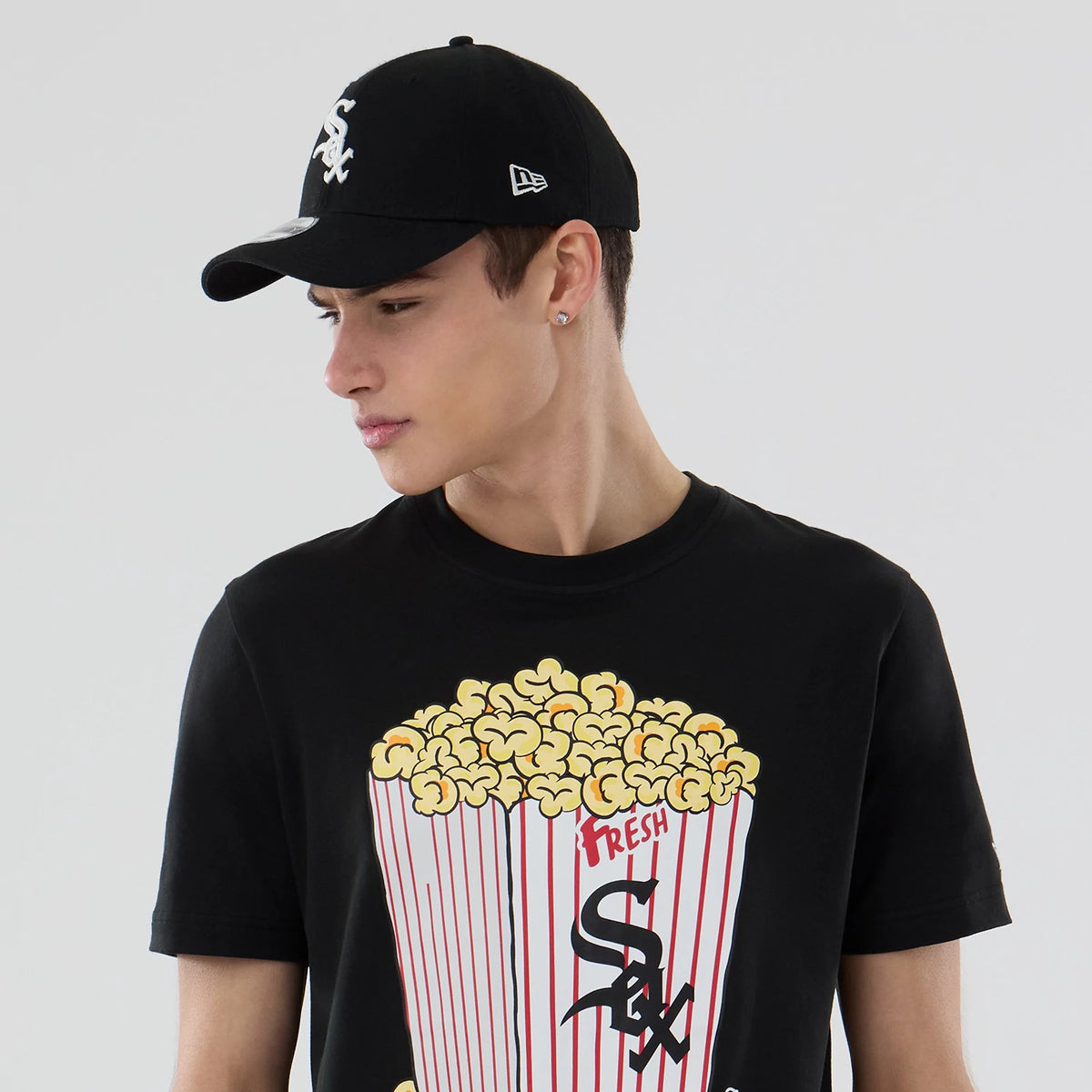 New Era Chicago White Sox MLB Popcorn Black T-Shirt, front view on-model showing the popcorn graphic with red and white stripes and Sox lettering across the chest, styled with a matching black cap featuring an embroidered logo on the front.