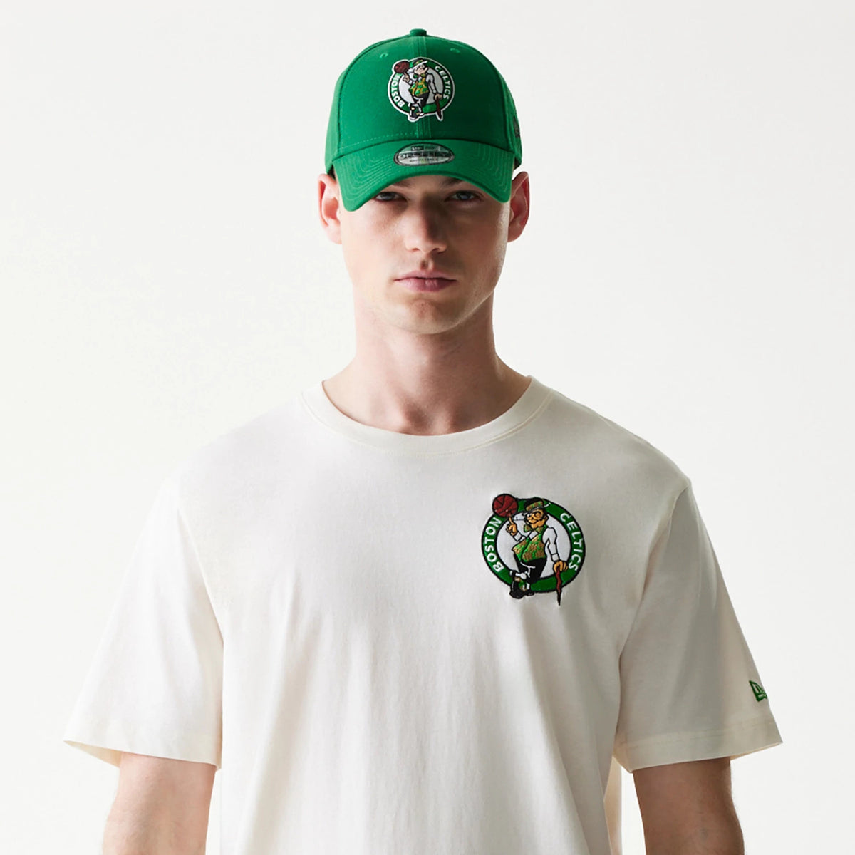New Era Boston Celtics NBA Hotdog Cream T-Shirt, front view on-model featuring an embroidered Celtics logo on the left chest, short sleeves, and a small New Era logo embroidered on the left sleeve, styled with a matching green cap.
