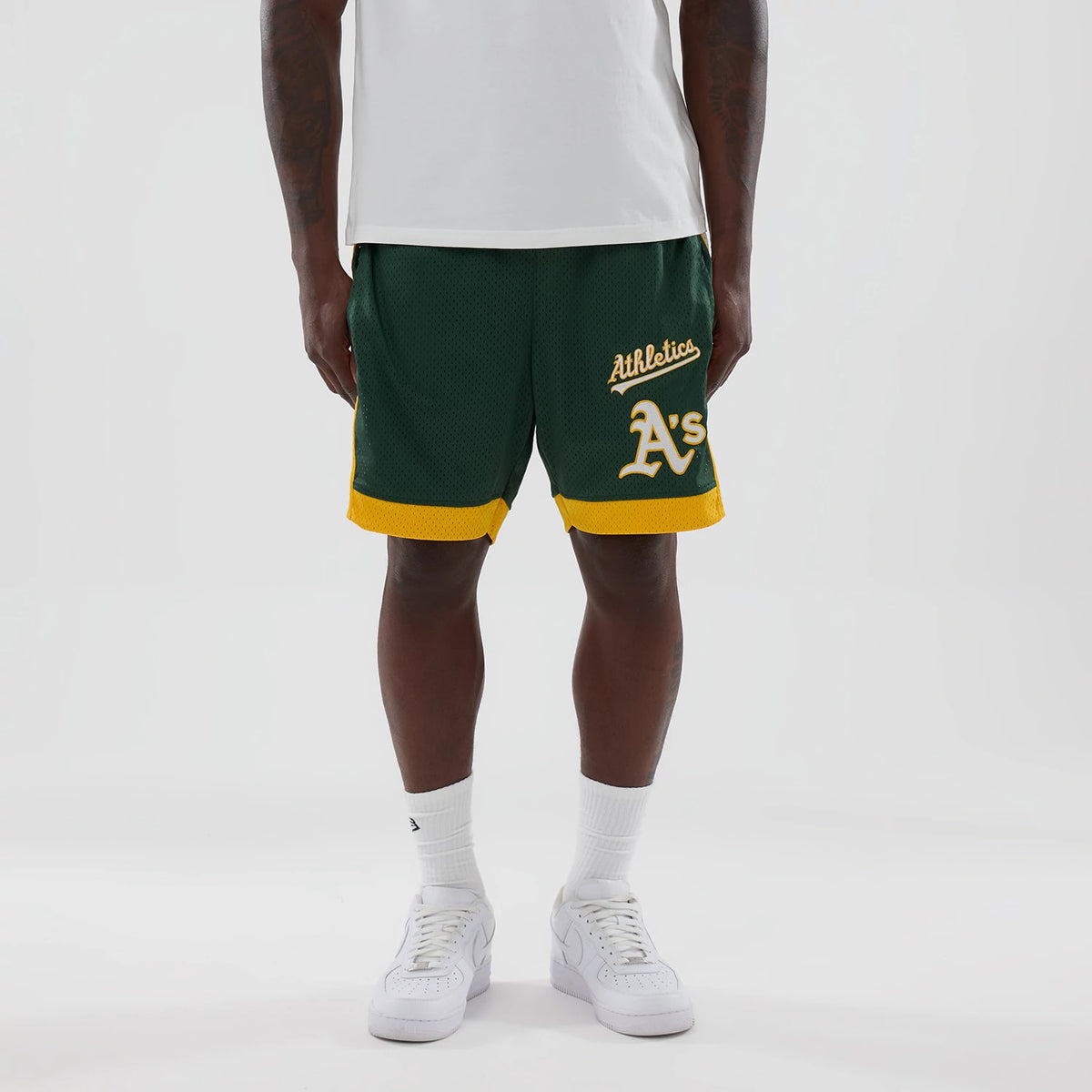 New Era Athletics MLB Dark Green Mesh Shorts, front view on a model with a white top, showing script Athletics lettering and a large A's logo on the right leg, accented by yellow trim around the hem.