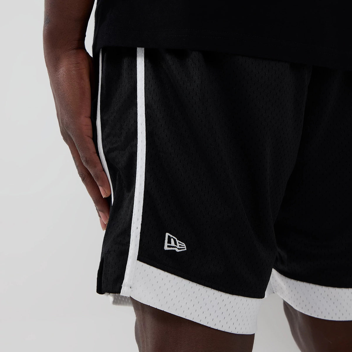 New Era Chicago Bulls NBA Black Mesh Shorts, side view showing black fabric with white trim along the seams and hem, an embroidered white New Era logo near the hem, worn on a model with a close-up of their hand.