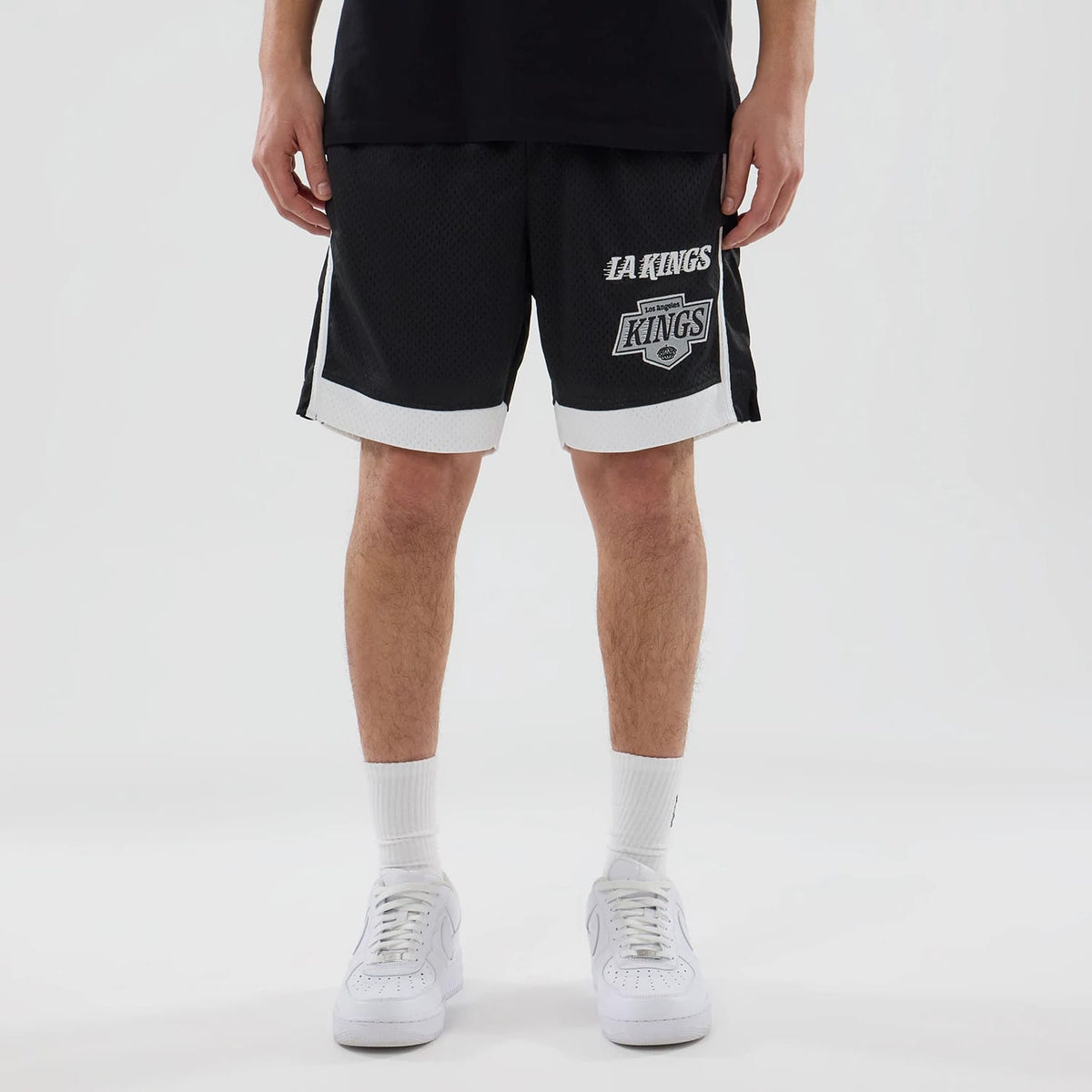 New Era LA Kings NHL Black Mesh Shorts, front view showing white printed LA Kings logo on the lower left leg, contrasting white trim on the hem and sides, styled with white socks and trainers on a model.