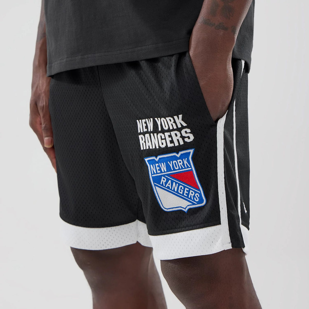 New Era New York Rangers NHL Black Mesh Shorts, front view on-model showing the white embroidered New York Rangers text and logo on the left leg, contrasted with a black mesh fabric and a white trim at the hem.