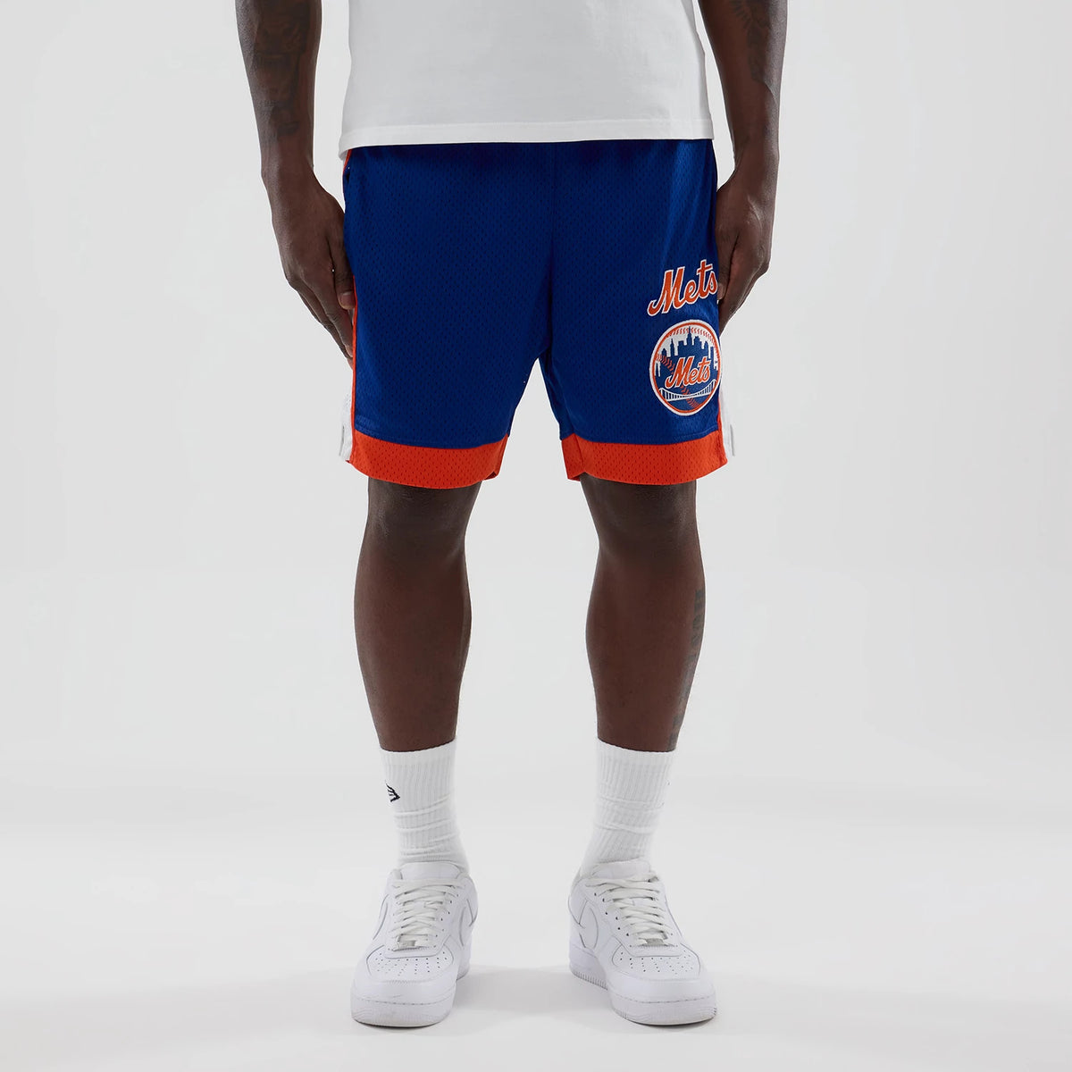 New Era New York Mets MLB Blue Mesh Shorts, front view on-model showing the blue mesh fabric with orange trim, an embroidered Mets logo on the left leg, and the 'Mets' script logo above it, styled with a white top and trainers.