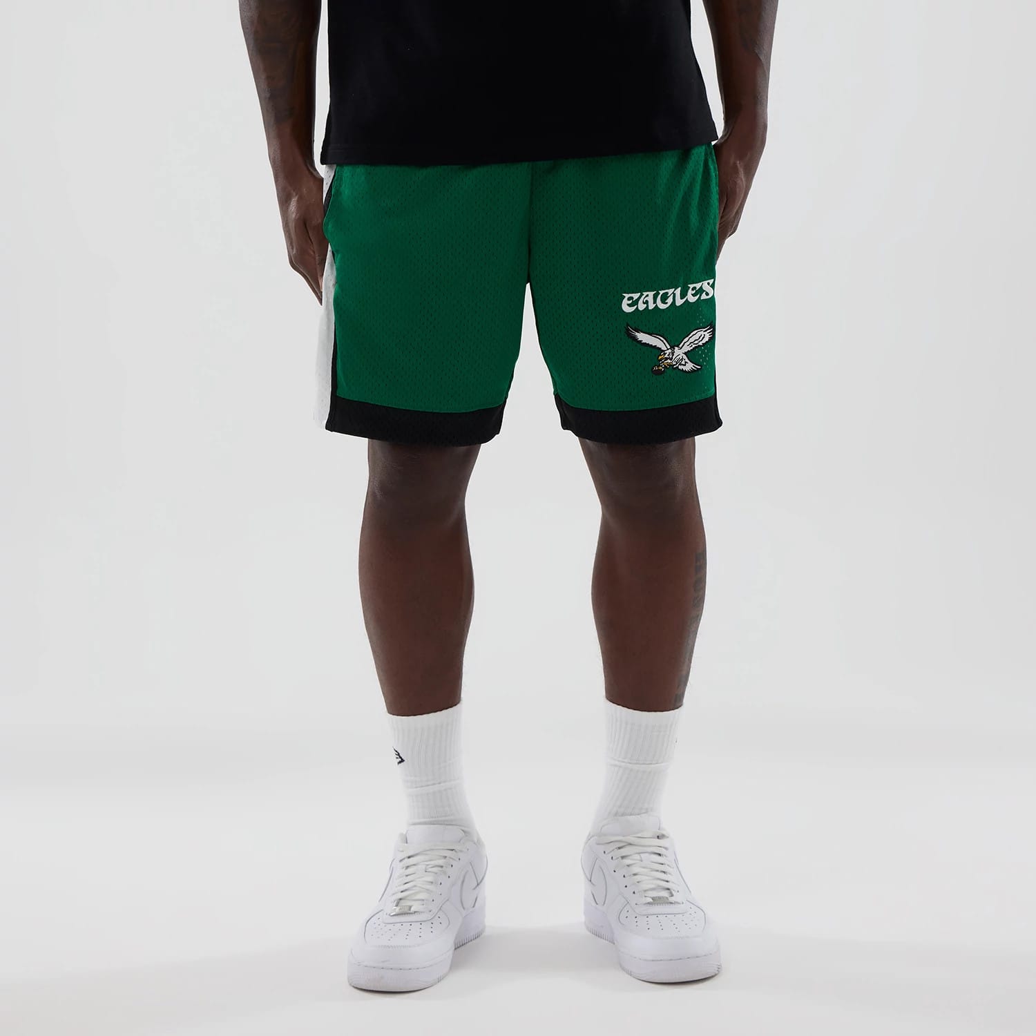 New Era Philadelphia Eagles NFL Green Mesh Shorts, front view on-model showing green mesh shorts with white Eagles lettering and an embroidered eagle logo on one leg, paired with a plain black top and white trainers.