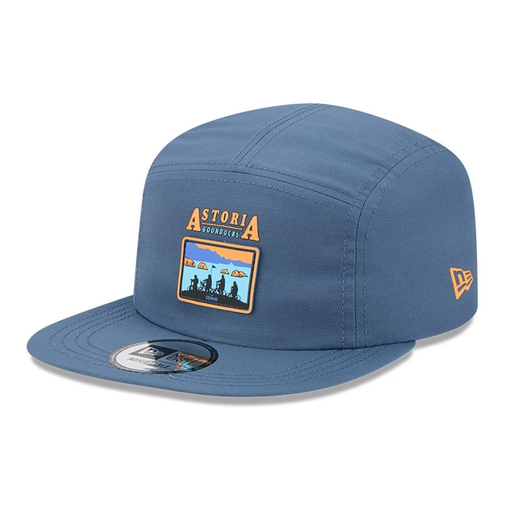 New Era The Goonies Astoria Warner Brothers Open Blue Camper Cap, three-quarter view showing the embroidered Astoria Good Docks patch on the front panel, flat blue visor with a 9FORTY size sticker and orange New Era flag logo on the left side.