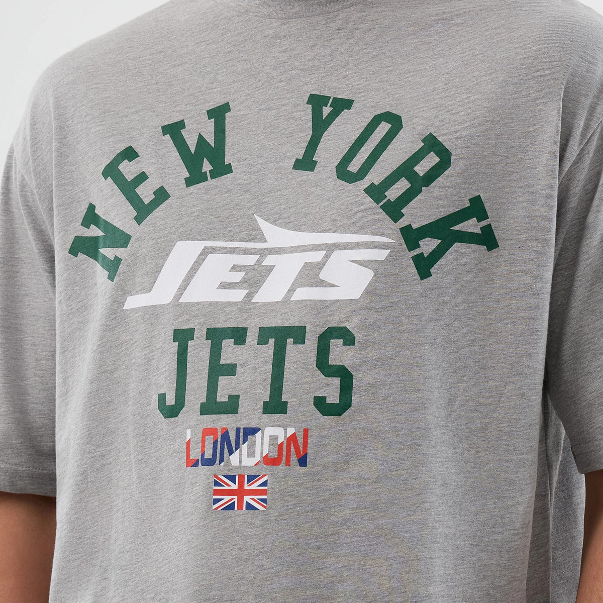 New Era New York Jets NFL International Series Games Grey Oversized T-Shirt, front view showing green New York lettering, a white Jets logo, green Jets text, red and blue London script, and a Union Jack below on the grey fabric.