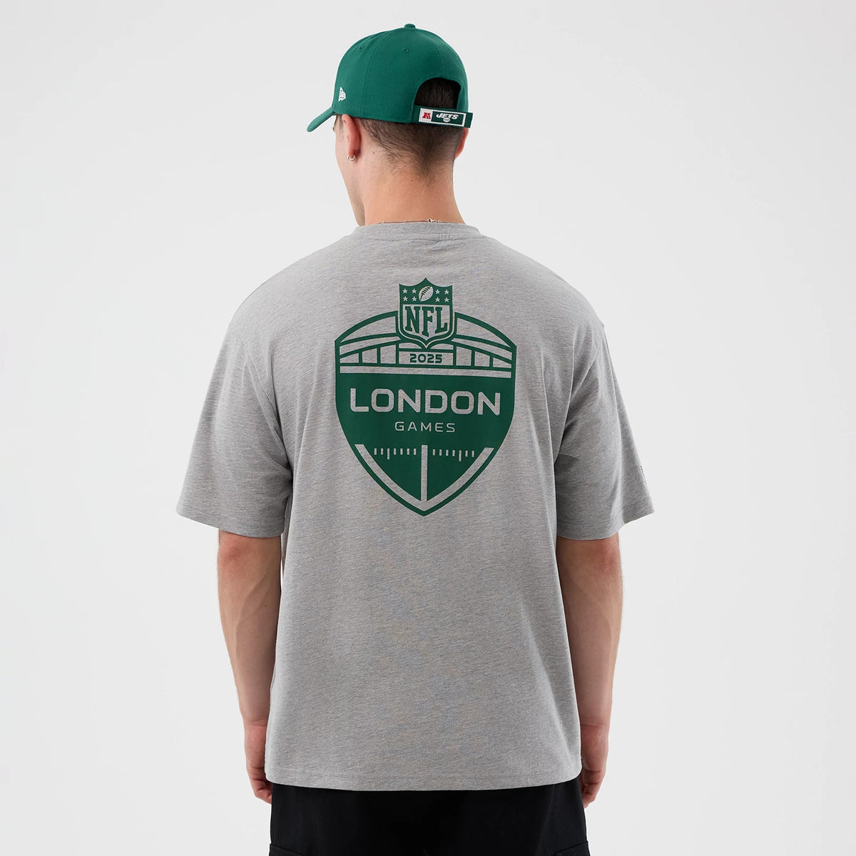 New Era New York Jets NFL International Series Games Grey Oversized T-Shirt, back view on a model with a matching green cap, featuring a large green London Games graphic centred on the upper back of the grey fabric.