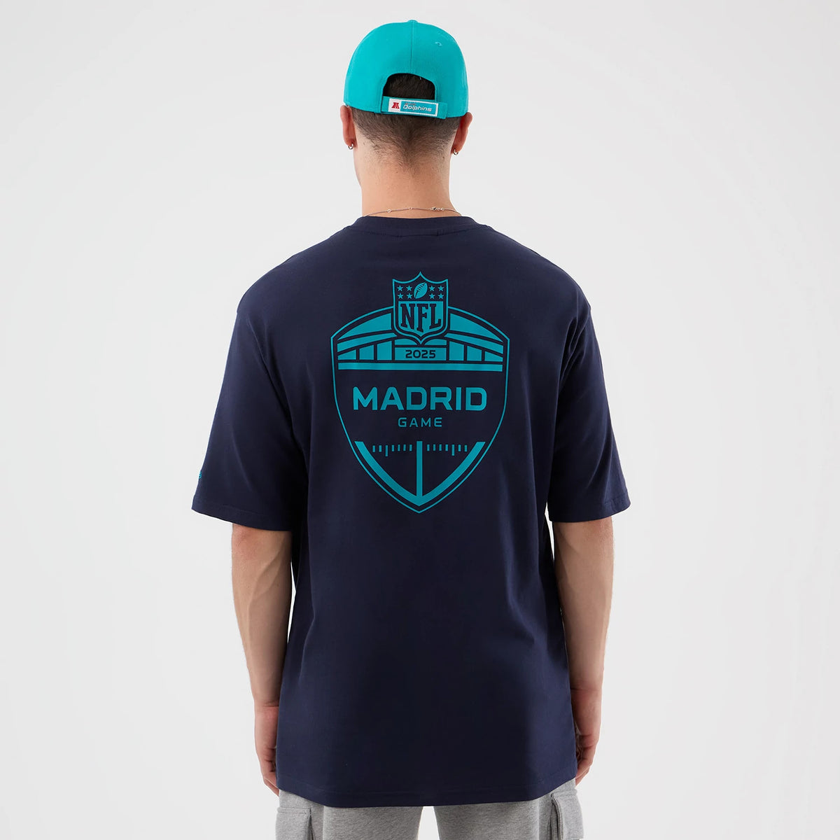 New Era Miami Dolphins NFL International Series Games Navy Oversized T-Shirt, back view on a model in a turquoise cap showing a large teal printed graphic with Madrid Game text and NFL 2023 emblem across the shoulders.