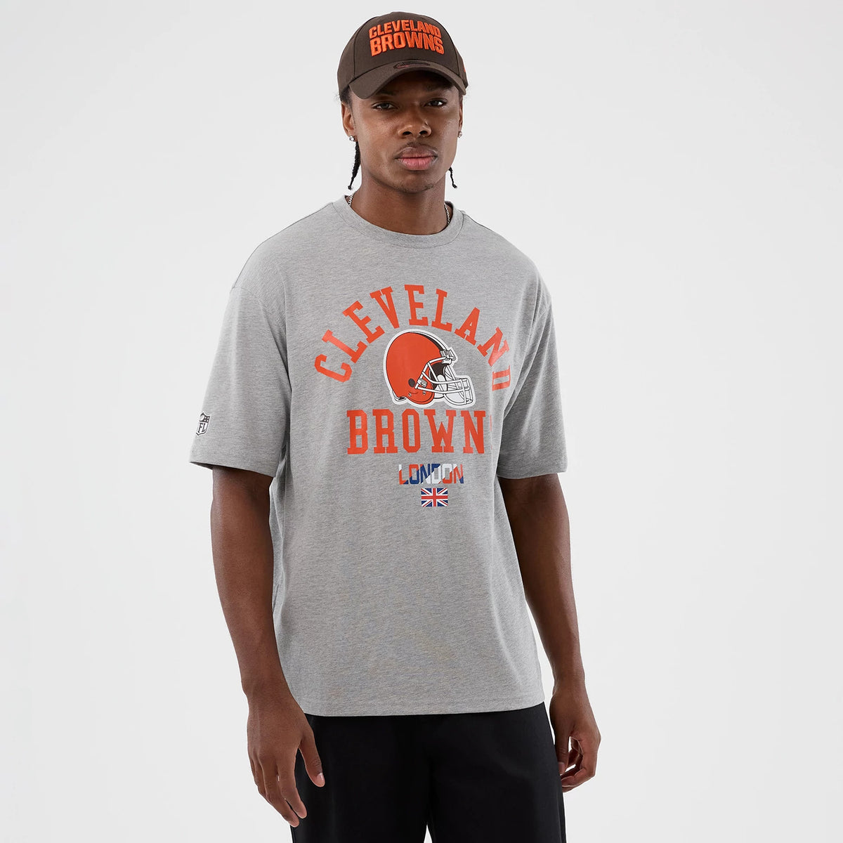 New Era Cleveland Browns NFL International Series Games Grey Oversized T-Shirt, front view on a model with a matching cap, featuring the Cleveland Browns helmet printed in orange with team name above, London below.