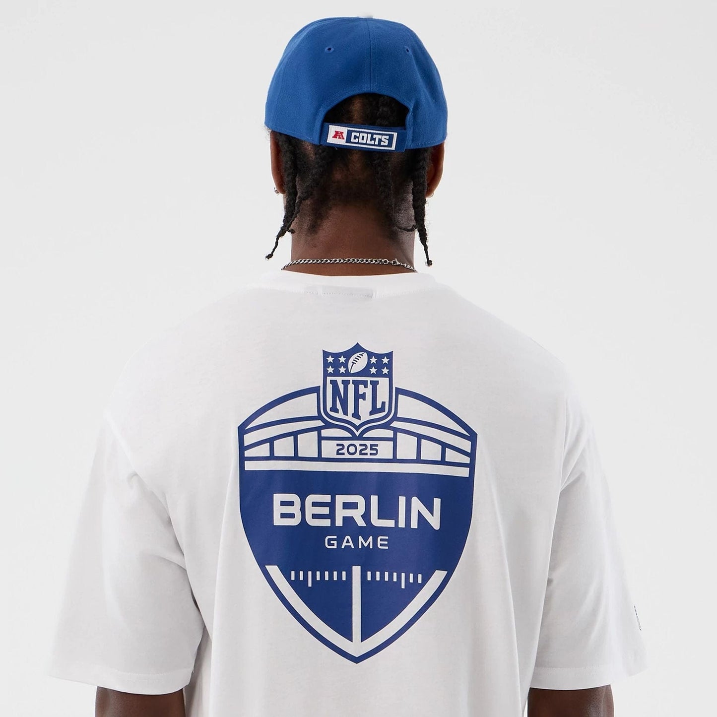 New Era Indianapolis Colts NFL International Series Games White Oversized T-Shirt, back view on a model showing a blue graphic with NFL 2025 and Berlin Game lettering, complemented by a blue cap with a Colts patch on the back.