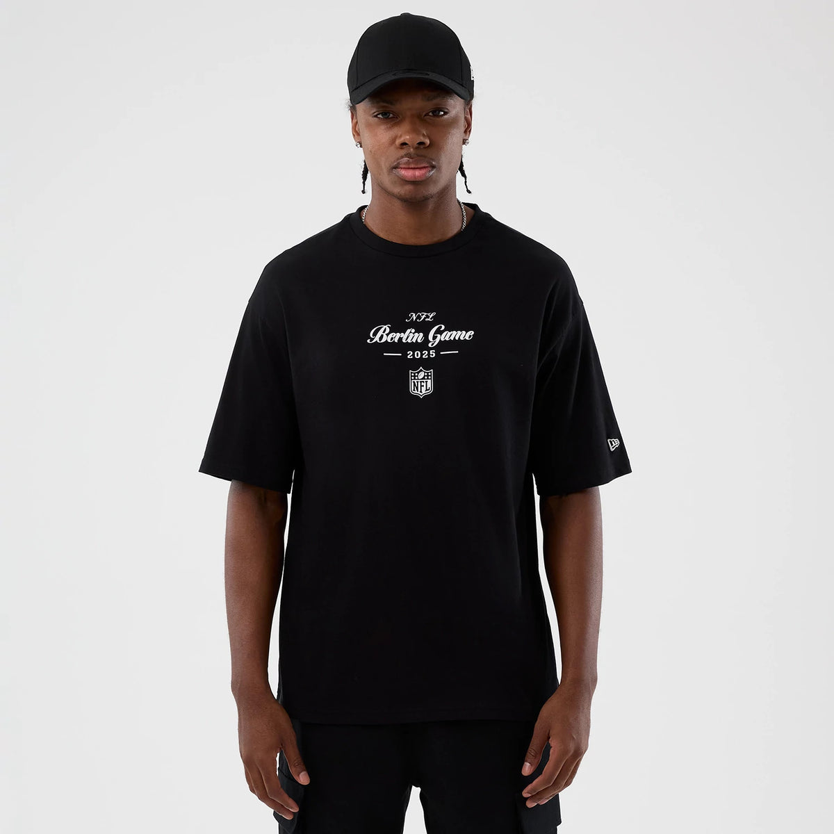 New Era Berlin NFL International Series Games Black Oversized T-Shirt, front view on a model paired with a plain black cap, featuring a central white printed Berlin Game 2023 graphic and a small embroidered New Era logo on the left sleeve.
