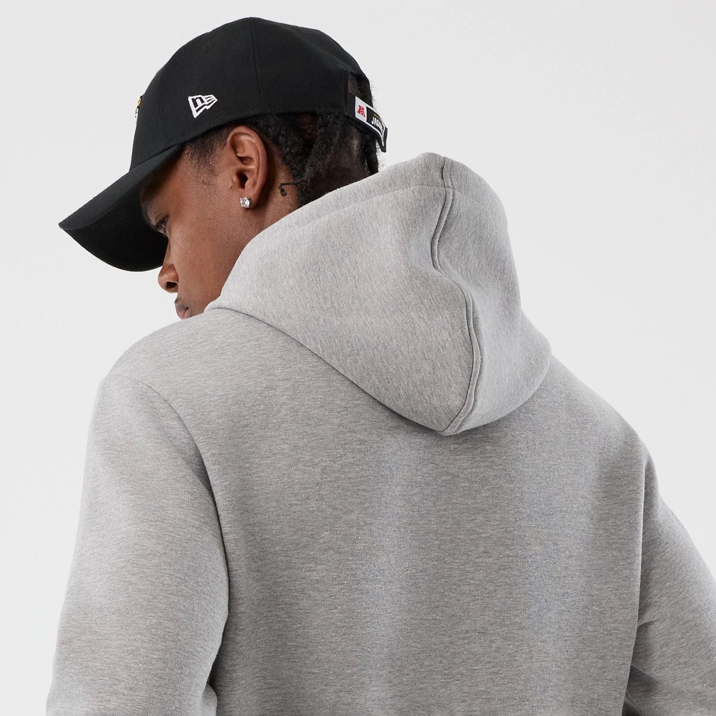 New Era Jacksonville Jaguars NFL International Series Games Grey Pullover Hoodie, back view on a model with a black cap featuring an embroidered New Era flag on the side, showcasing the simple grey fabric and hood detail.