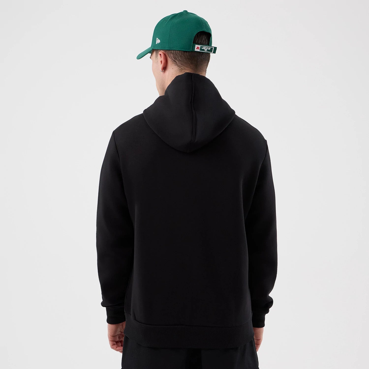 New Era New York Jets NFL International Series Games Black Pullover Hoodie, back view on-model showing the plain black back with a spacious hood, styled casually with a contrasting green cap featuring an embroidered New Era logo on the side.