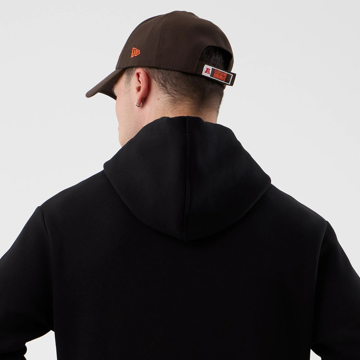 New Era Cleveland Browns NFL International Series Games Black Pullover Hoodie, back view on-model showing the plain black hood with a brown cap featuring a debossed orange New Era logo on the side.