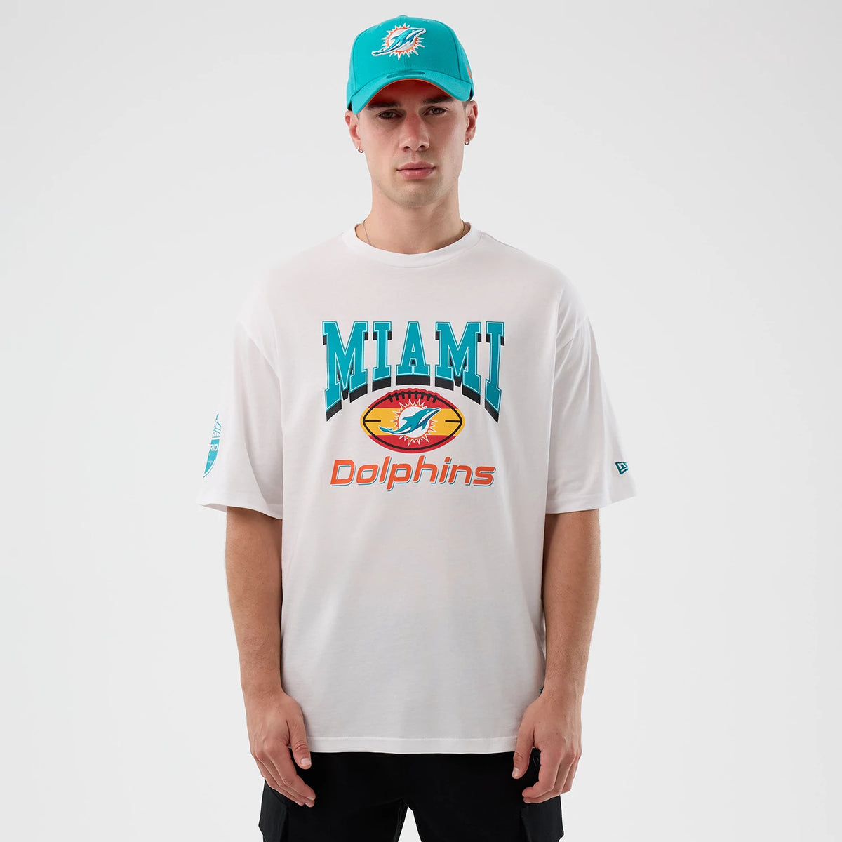 New Era Dolphins NFL International Series Games Madrid White Oversized T-Shirt, front view on a model with a teal cap showing Miami Dolphins graphics across the chest and a small embroidered New Era logo on the left sleeve.