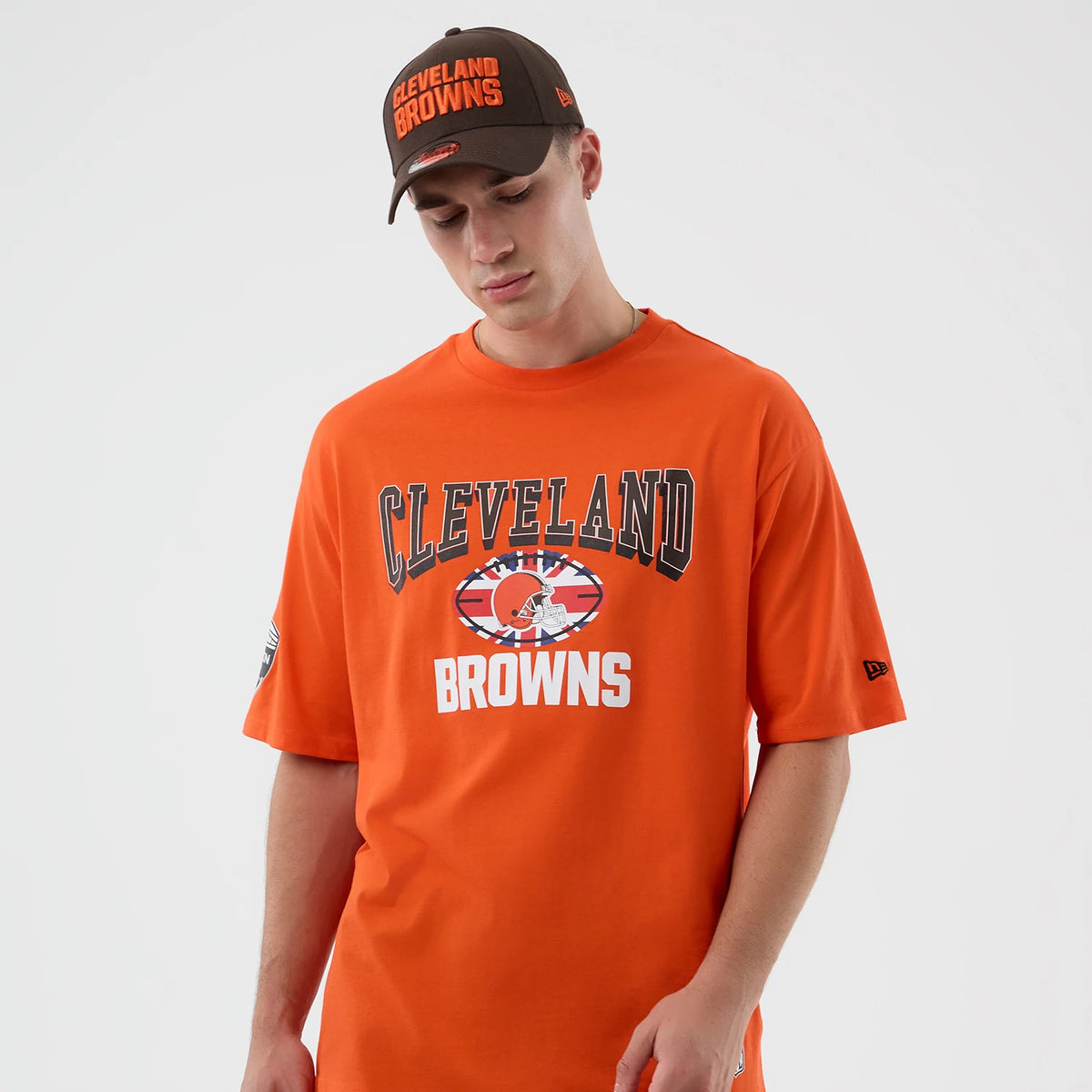 New Era Cleveland Browns NFL International Series Games Orange Oversized T-Shirt, front view on-model featuring Cleveland Browns lettering across the chest, a centred team logo with a flag design, and a small embroidered New Era logo on the sleeve.