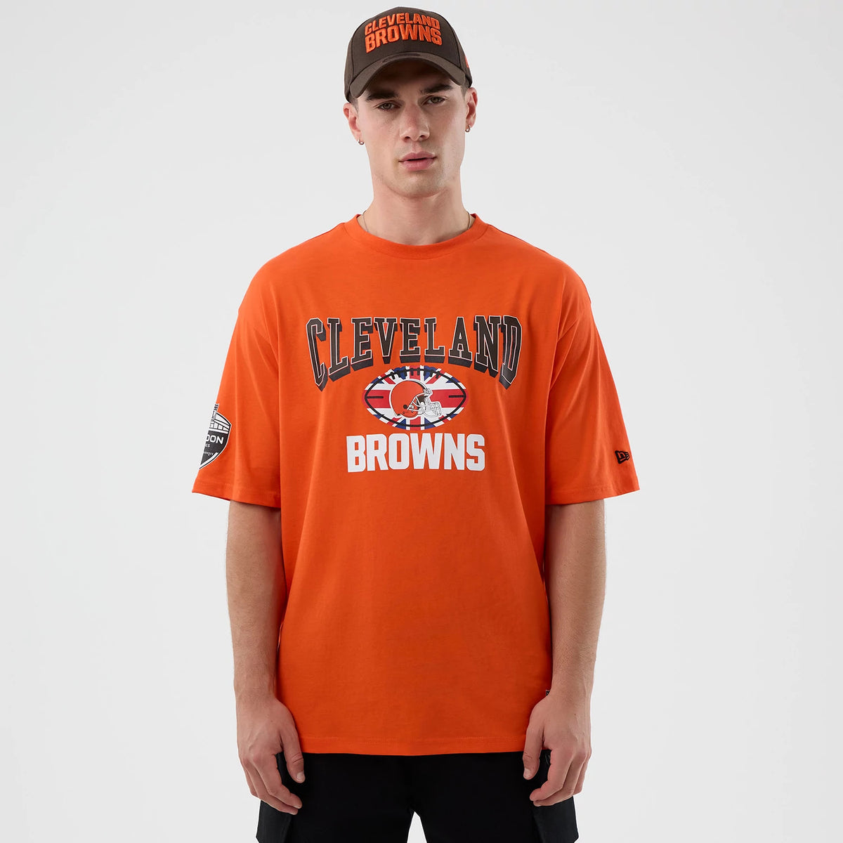 New Era Cleveland Browns NFL International Series Games Orange Oversized T-Shirt, front view on a model with a matching cap, showing Cleveland Browns graphics across the chest and an embroidered New Era logo on the left sleeve.