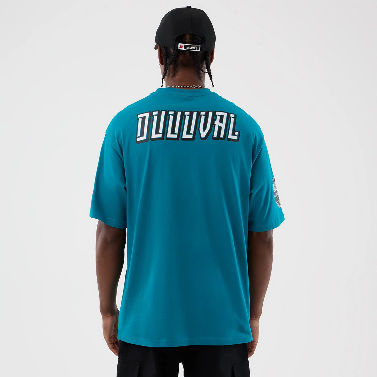 New Era Jacksonville Jaguars NFL International Series Games Turquoise Oversized T-Shirt, back view on a model featuring Duval lettering across the upper back and an embroidered logo on the right sleeve, styled with a black cap.