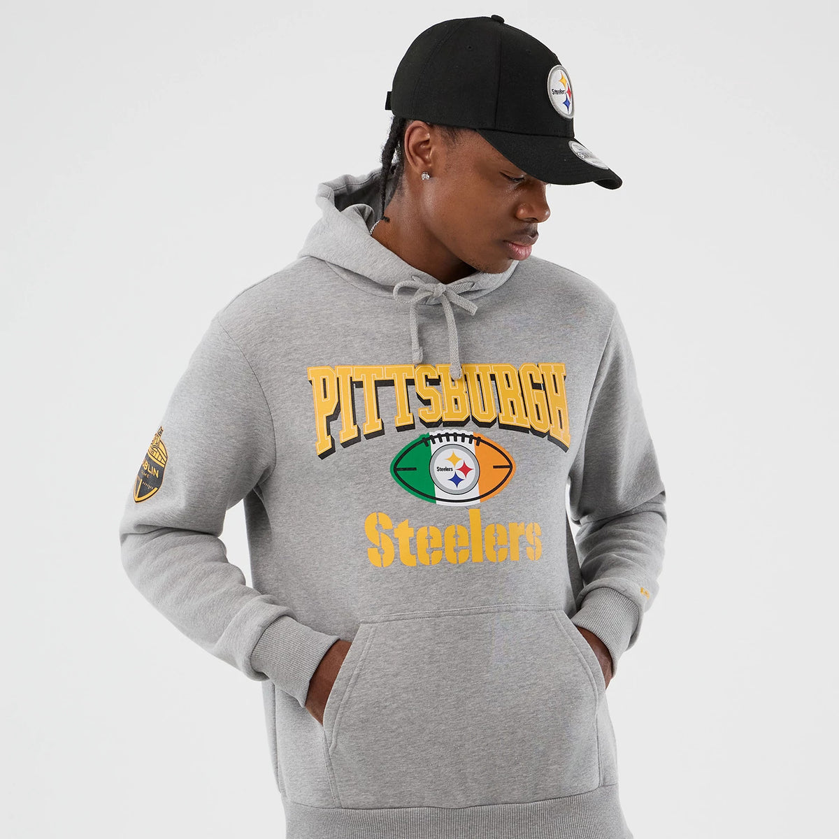 New Era Pittsburgh Steelers NFL International Series Games Grey Pullover Hoodie, front view on a model with black cap showing large printed Pittsburgh Steelers lettering across the chest and a team logo football graphic, with sleeve patch detail.