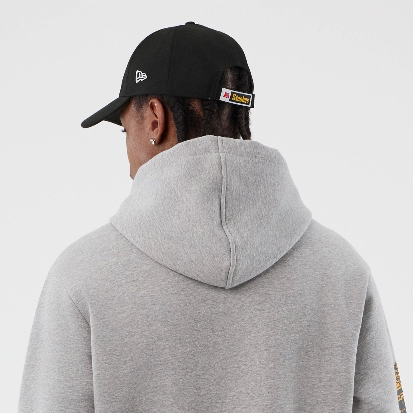 New Era Pittsburgh Steelers NFL International Series Games Grey Pullover Hoodie, back view on model showing the plain grey hood and shoulders, with a black cap featuring an embroidered New Era logo and a Steelers tab at the back.