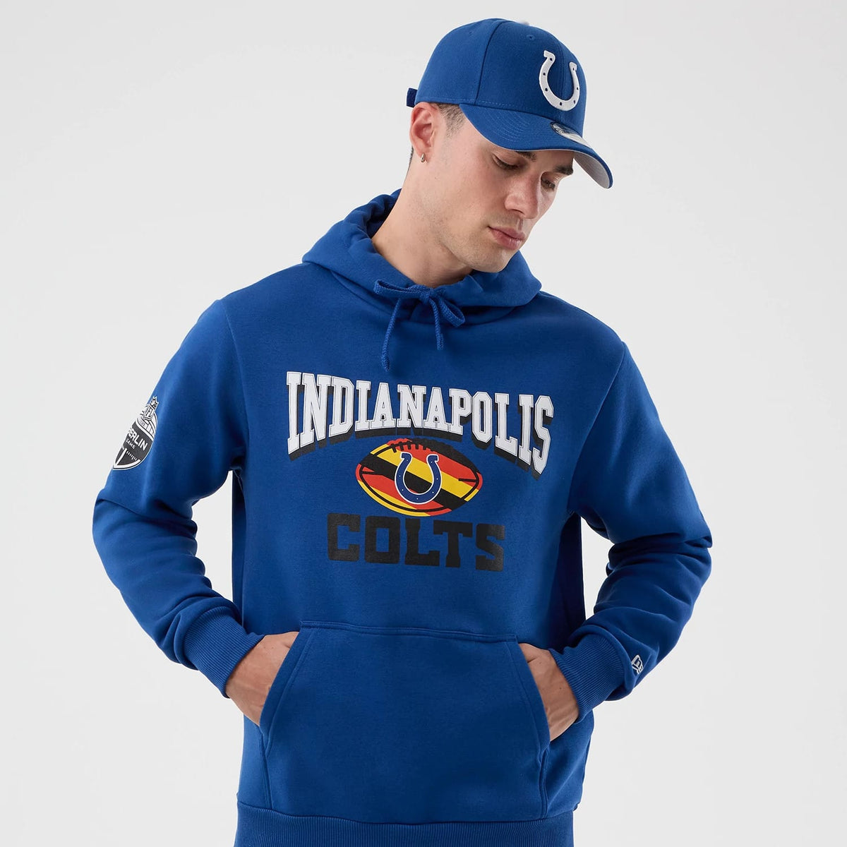 New Era Indianapolis Colts NFL International Series Games Blue Pullover Hoodie, front view on a model with matching blue cap, featuring a printed Indianapolis Colts logo centred on the chest and an embroidered New Era logo on the cuff.