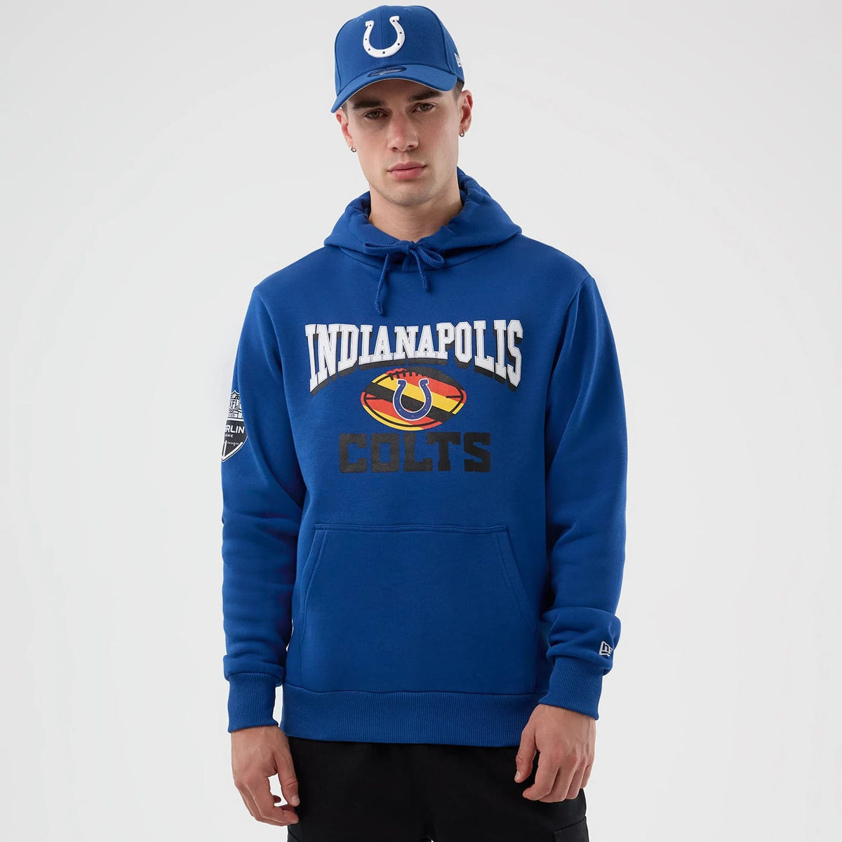 New Era Indianapolis Colts NFL International Series Games Blue Pullover Hoodie, front view on-model showing printed Colts lettering and logo on chest, NFL shield on the sleeve, and embroidered New Era logo near the cuff, styled with a matching cap.