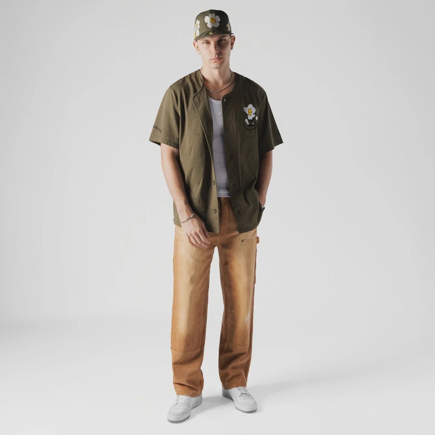 New Era x Rodrigo Roji Flower Green Jersey, on-model front view with white and yellow flower graphics on the cap and left chest of the green jersey, styled with tan trousers and white sneakers.