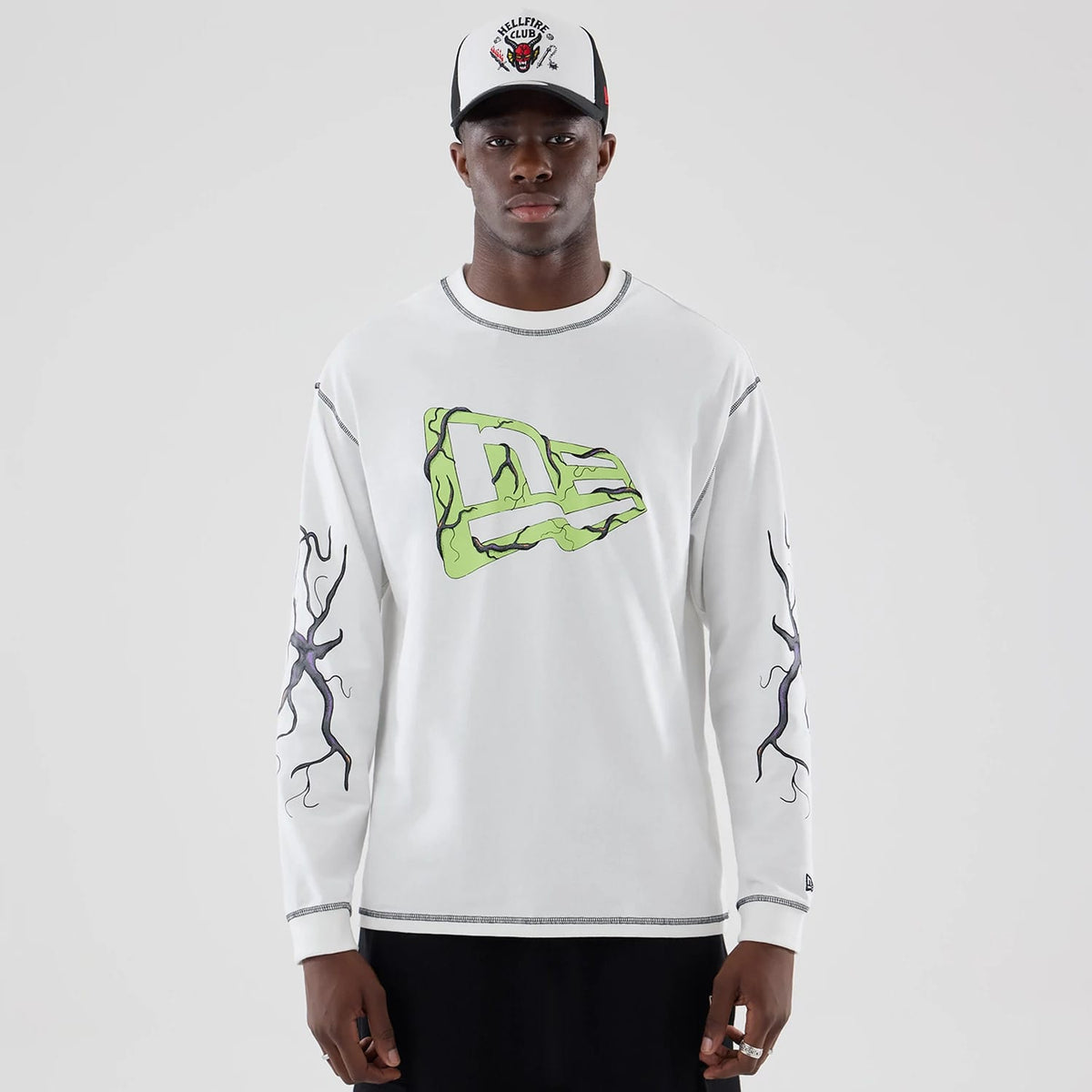 New Era Stranger Things White Oversized Long Sleeve T-Shirt, front view on-model featuring a large green and black graphic New Era logo across the chest and intricate black vine designs running down both sleeves.
