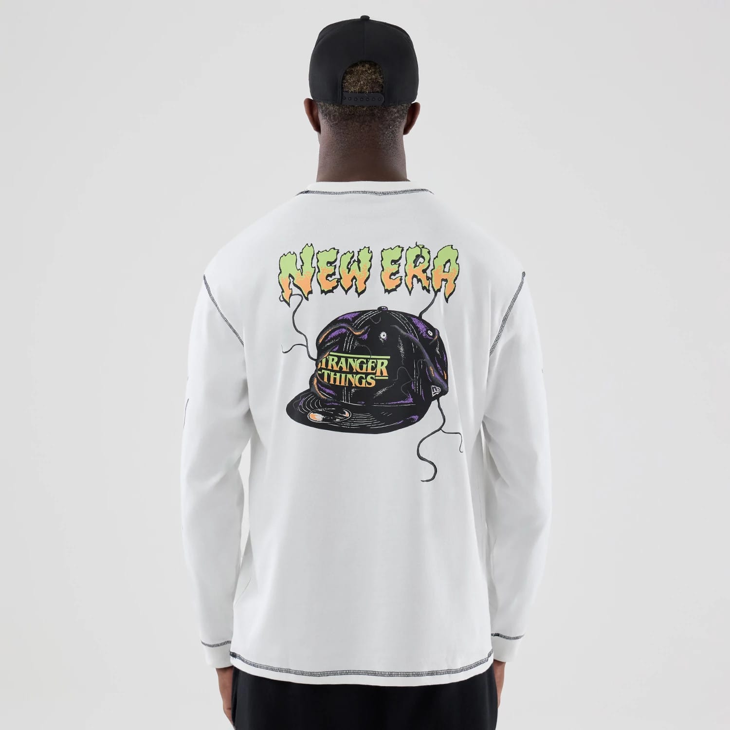 New Era Stranger Things White Oversized Long Sleeve T-Shirt, back view showing a large graphic of a black cap with Stranger Things text and colourful New Era lettering above, worn by a model with black trousers and a black cap.