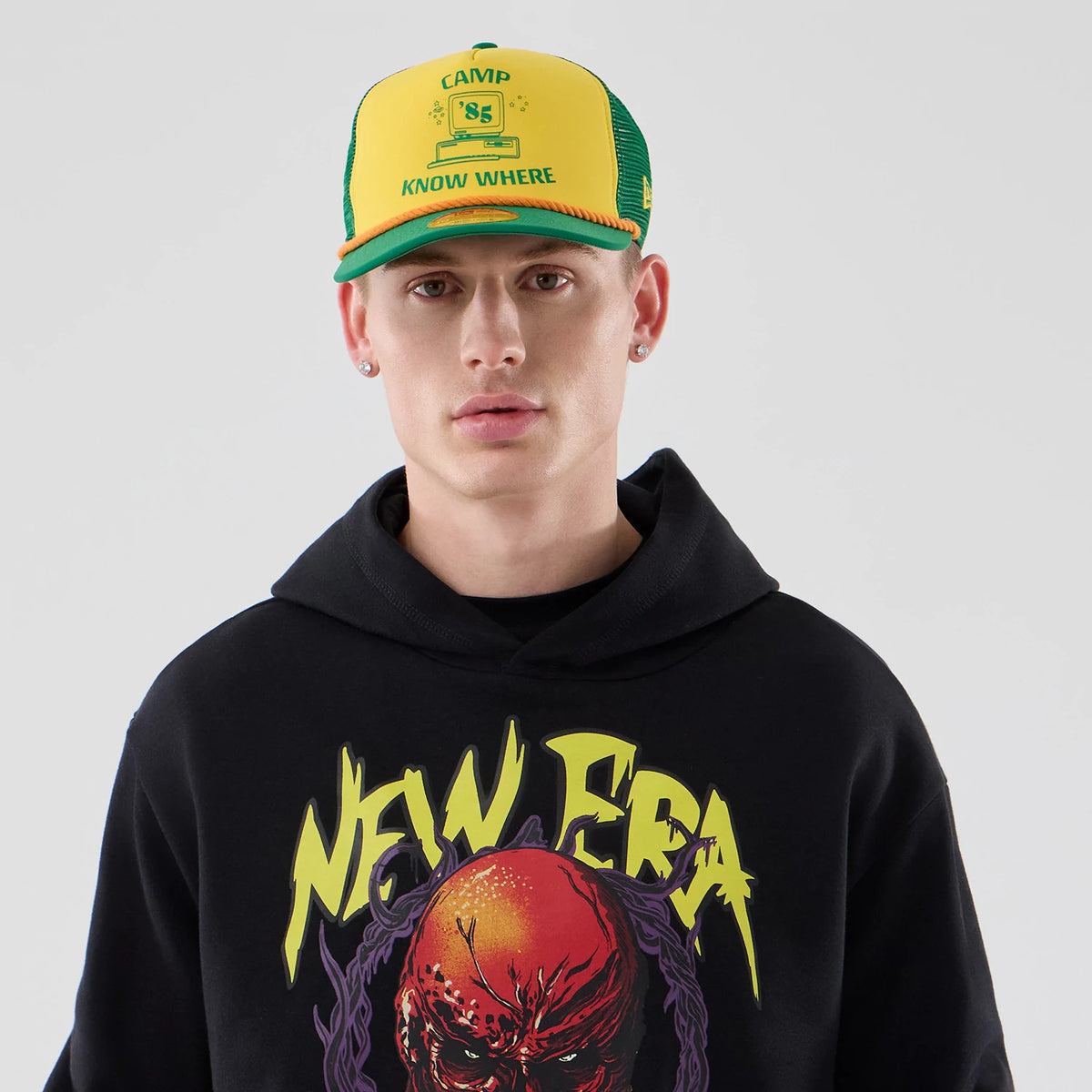 New Era Stranger Things Black Oversized Pullover Hoodie, front view on-model showcasing the graphic on the chest with New Era branding, styled with a yellow and green cap featuring Camp Know Where lettering on the front panel.