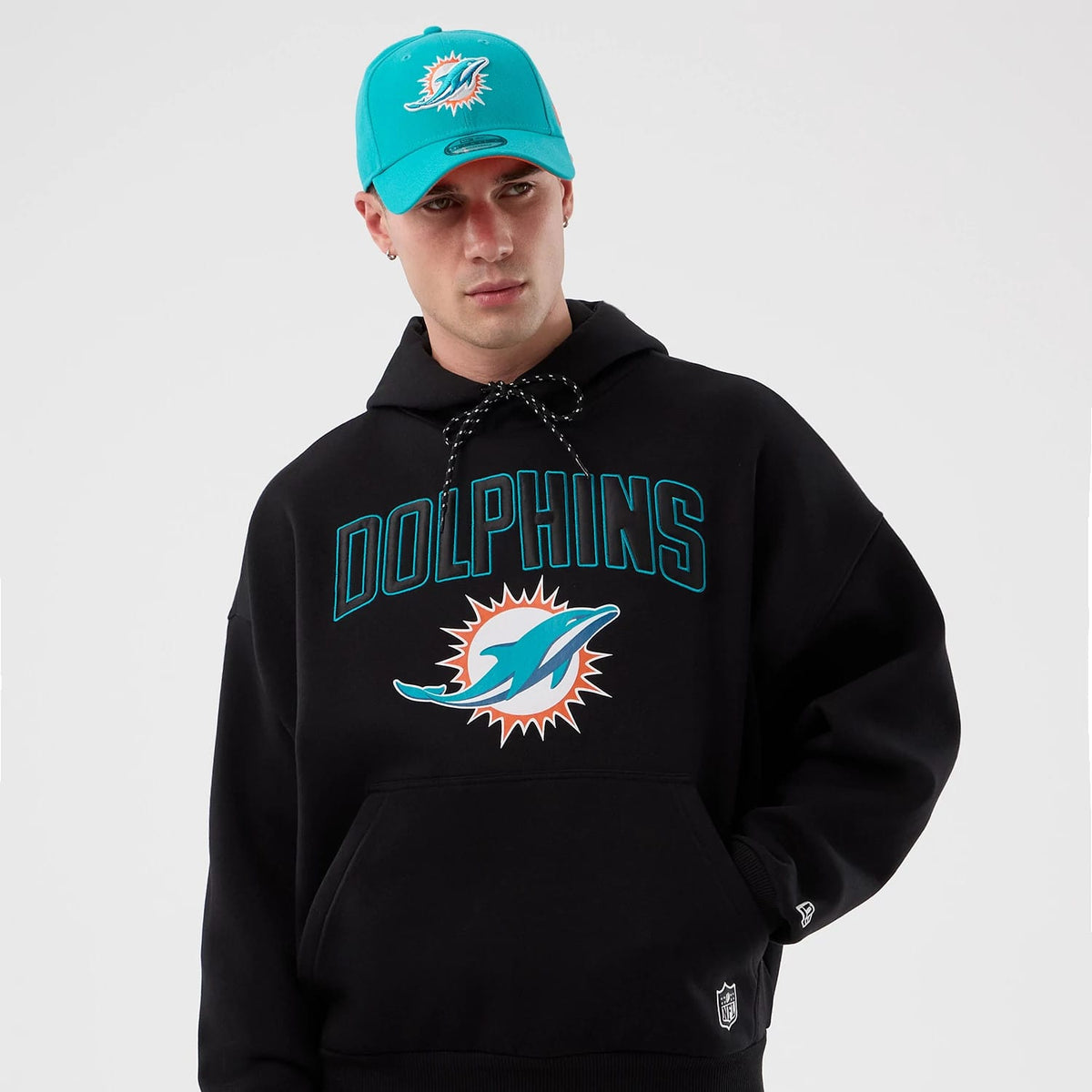 New Era Miami Dolphins NFL International Series Games Black Pullover Hoodie, front view on a model pairing with a turquoise cap, featuring an embroidered Dolphins logo and lettering across the chest with a drawstring hood.