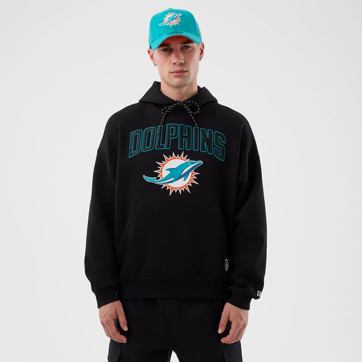 New Era Miami Dolphins NFL International Series Games Black Pullover Hoodie, front view on a model wearing a matching cap, featuring large teal Dolphins lettering with a logo on the chest and a small embroidered New Era logo near the cuff.