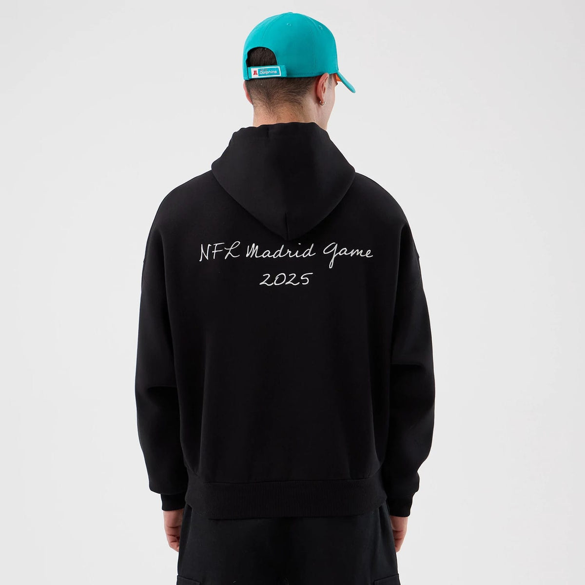 New Era Miami Dolphins NFL International Series Games Black Pullover Hoodie, back view showing NFL Madrid Game 2025 lettering across the upper back, with the hood resting flat, on-model styled with a teal cap.
