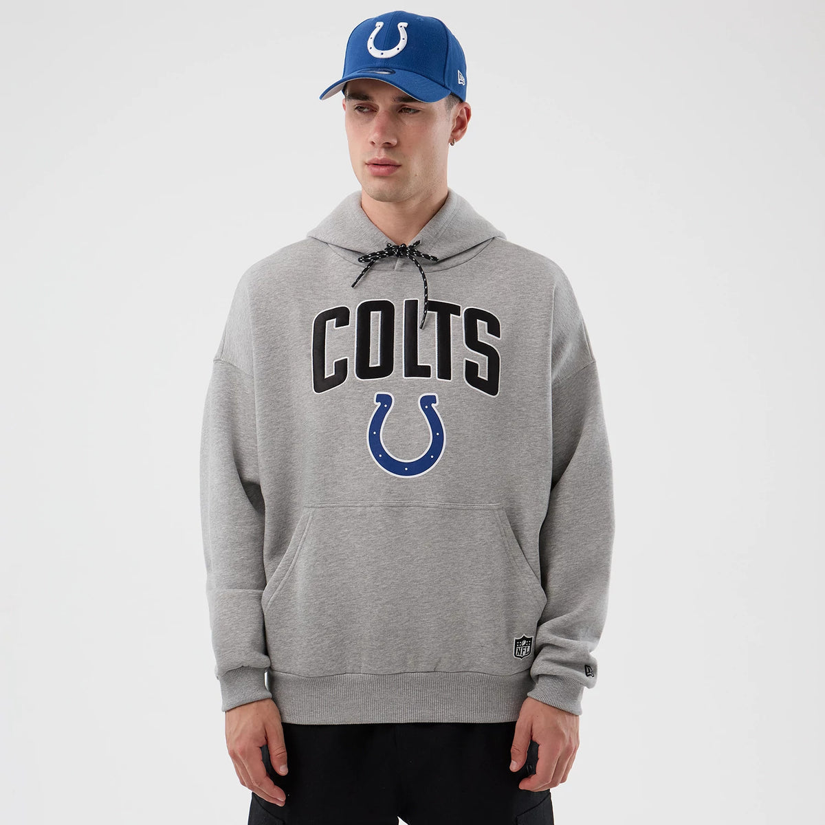 New Era Indianapolis Colts NFL International Series Games Grey Pullover Hoodie, front view on-model showing large COLTS lettering in black across the chest and a blue horseshoe below, with a small embroidered logo on the lower left side.