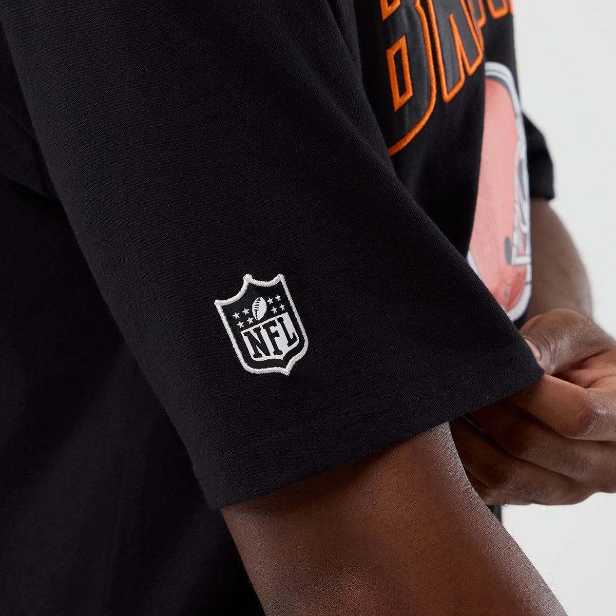 New Era Cleveland Browns NFL International Series Games Black Oversized T-Shirt, three-quarter view showing an embroidered white NFL logo on the sleeve and a close-up of the large orange and white team graphic on the chest.