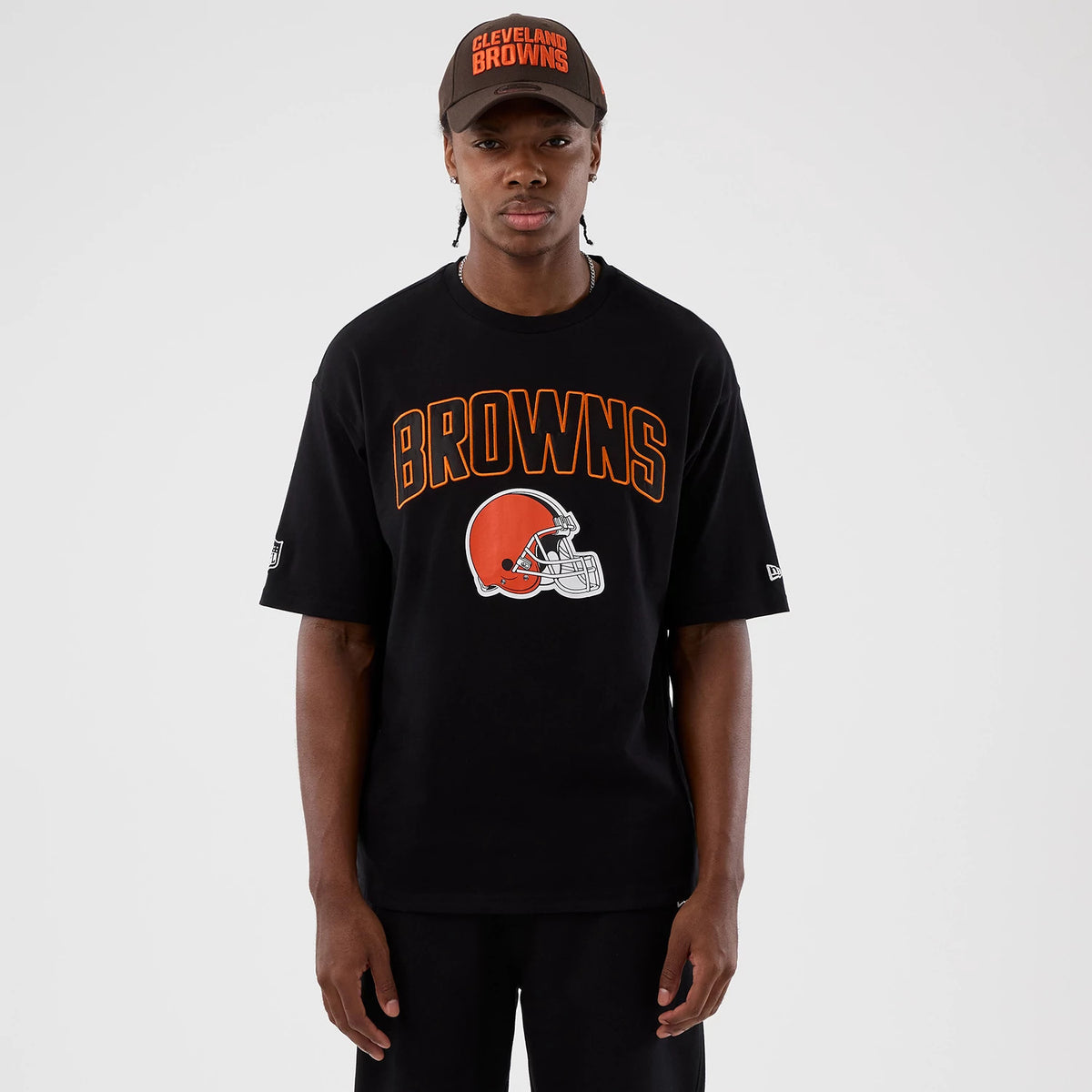 New Era Cleveland Browns NFL International Series Games Black Oversized T-Shirt, front view on a model with matching cap, displaying brown lettering above an orange Cleveland Browns helmet graphic, an embroidered New Era logo on the sleeve.