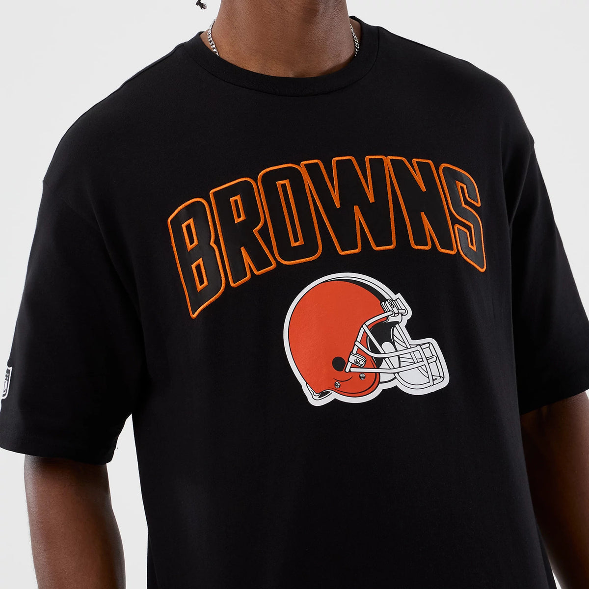 New Era Cleveland Browns NFL International Series Games Black Oversized T-Shirt, front view on-model showing orange BROWNS lettering and a printed orange helmet graphic centred on the chest, with a New Era logo patch on the left sleeve.
