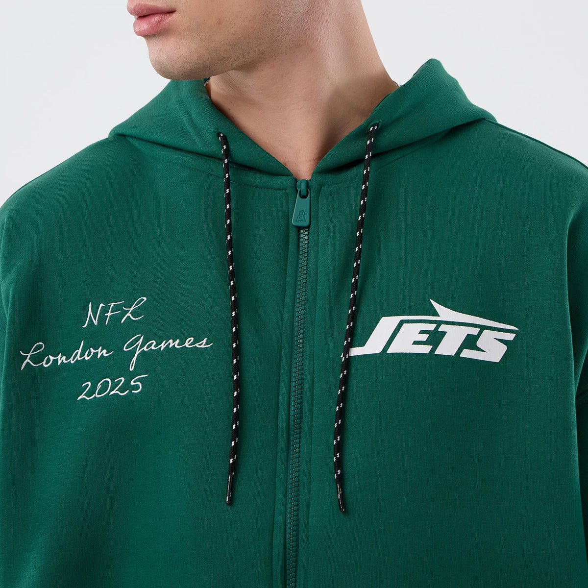 New Era New York Jets NFL International Series Games Green Full Zip Hoodie, front view showing white embroidered NFL London Games 2025 lettering on the left chest and a printed Jets logo on the right chest, with black-and-white striped drawstrings.