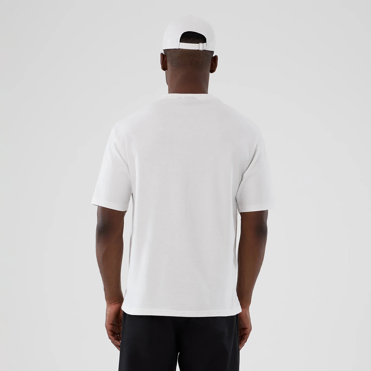 New Era Halloween Skull Graphic White Oversized T-Shirt, back view on a model showing a plain white back with short sleeves, paired with a white cap and dark trousers, highlighting the T-shirt's oversized fit and simple design.