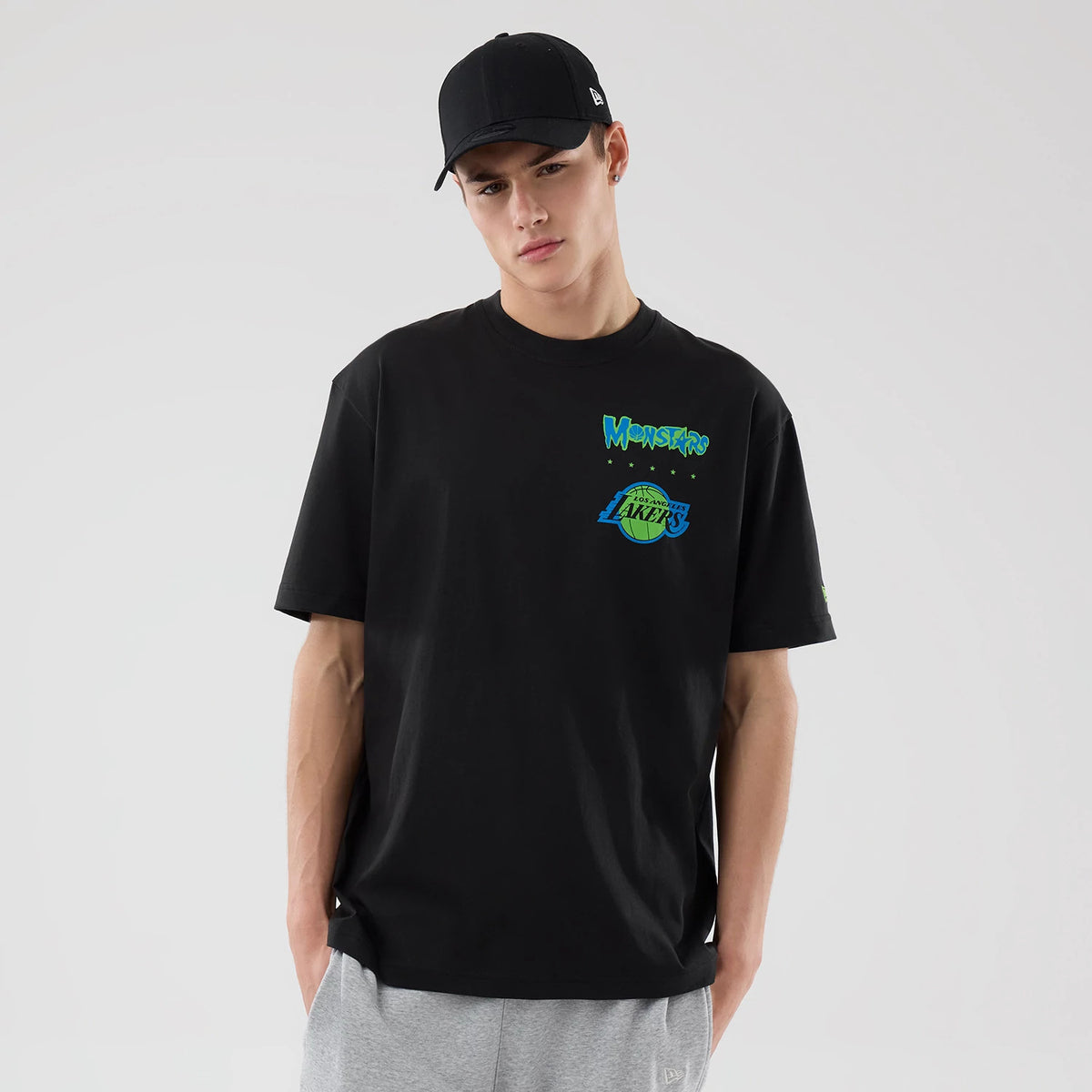 New Era LA Lakers Space Jam 2 X NBA Black T-Shirt, front view on a model with a black cap, showing the Monsters graphic and Lakers logo in green and blue on the chest, with plain short sleeves and a relaxed fit.