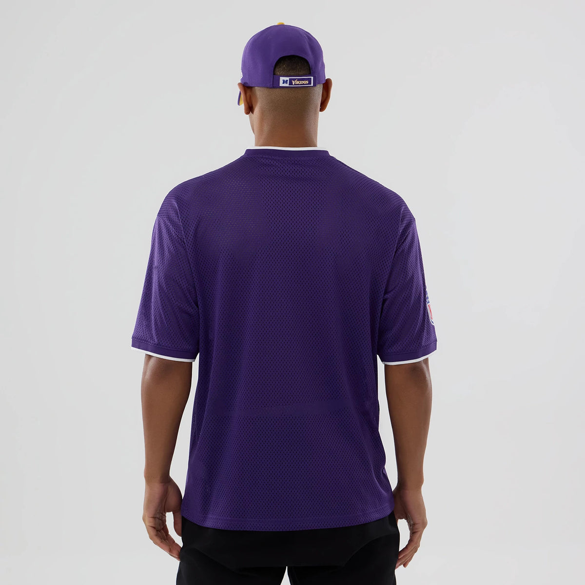 New Era Minnesota Vikings NFL Mesh Purple Oversized T-Shirt, back view on a model wearing a matching purple cap, showing the plain mesh design with white trim on the sleeves and collar, capturing a clean and simple look.