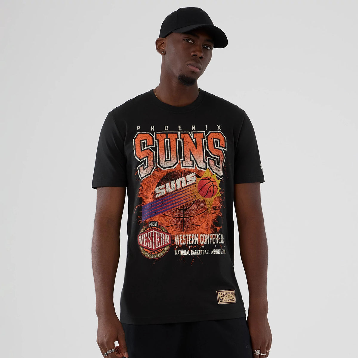 New Era Phoenix Suns NBA Hardwood Classic Black T-Shirt, front view on a model showing a graphic with large orange and grey SUNS text, a basketball design, and Western Conference lettering, with a patch near the bottom hem.