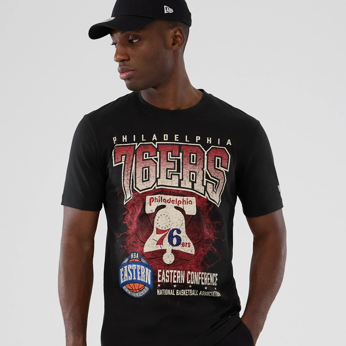 New Era Philadelphia 76ers NBA Hardwood Classic Black T-Shirt, front view on a model wearing a matching cap, featuring 76ers graphics in red and white on the chest and an embroidered New Era logo on the sleeve.