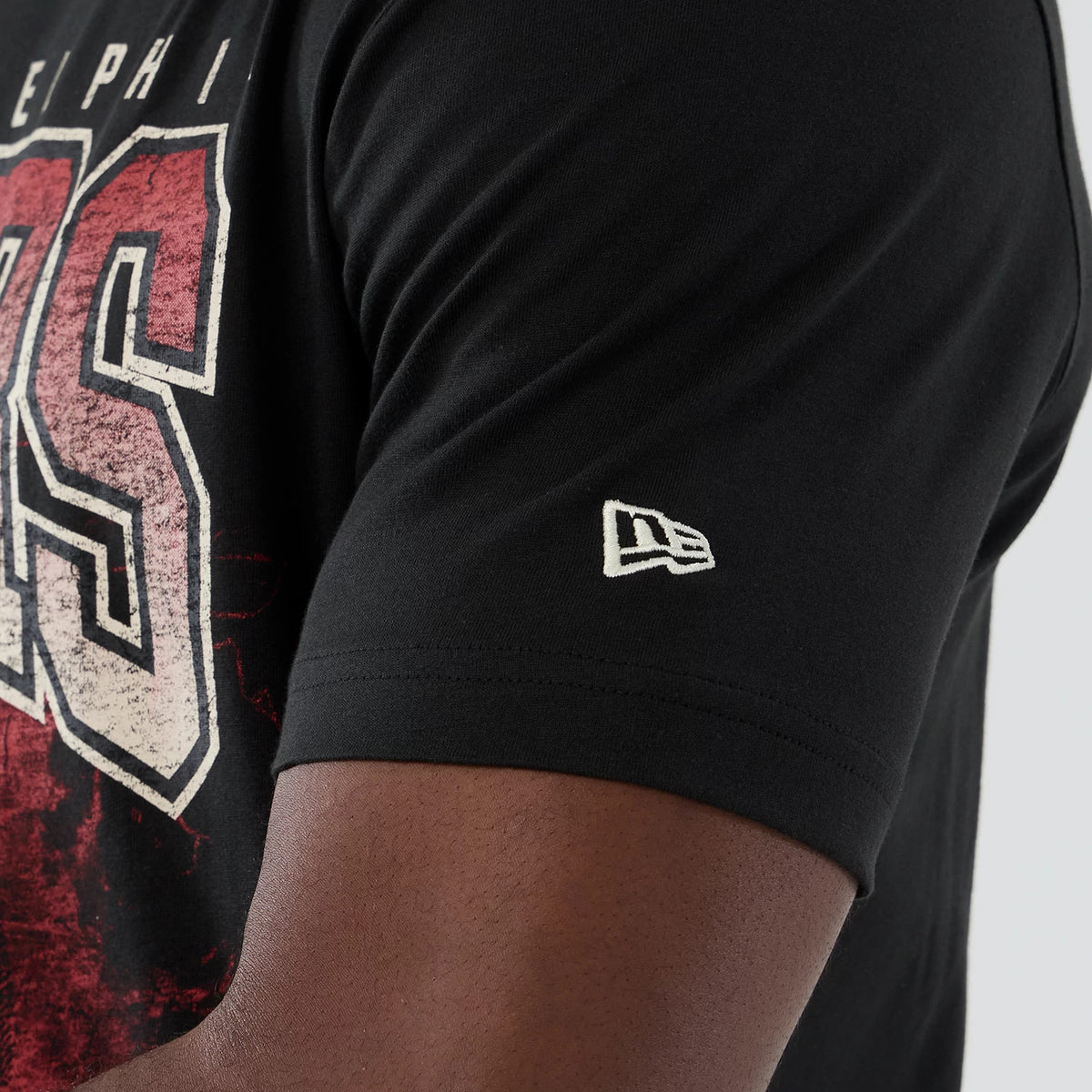 New Era Philadelphia 76ers NBA Hardwood Classic Black T-Shirt, side view showing the red and white 76ers graphic across the chest and an embroidered white New Era logo on the sleeve against the black fabric.