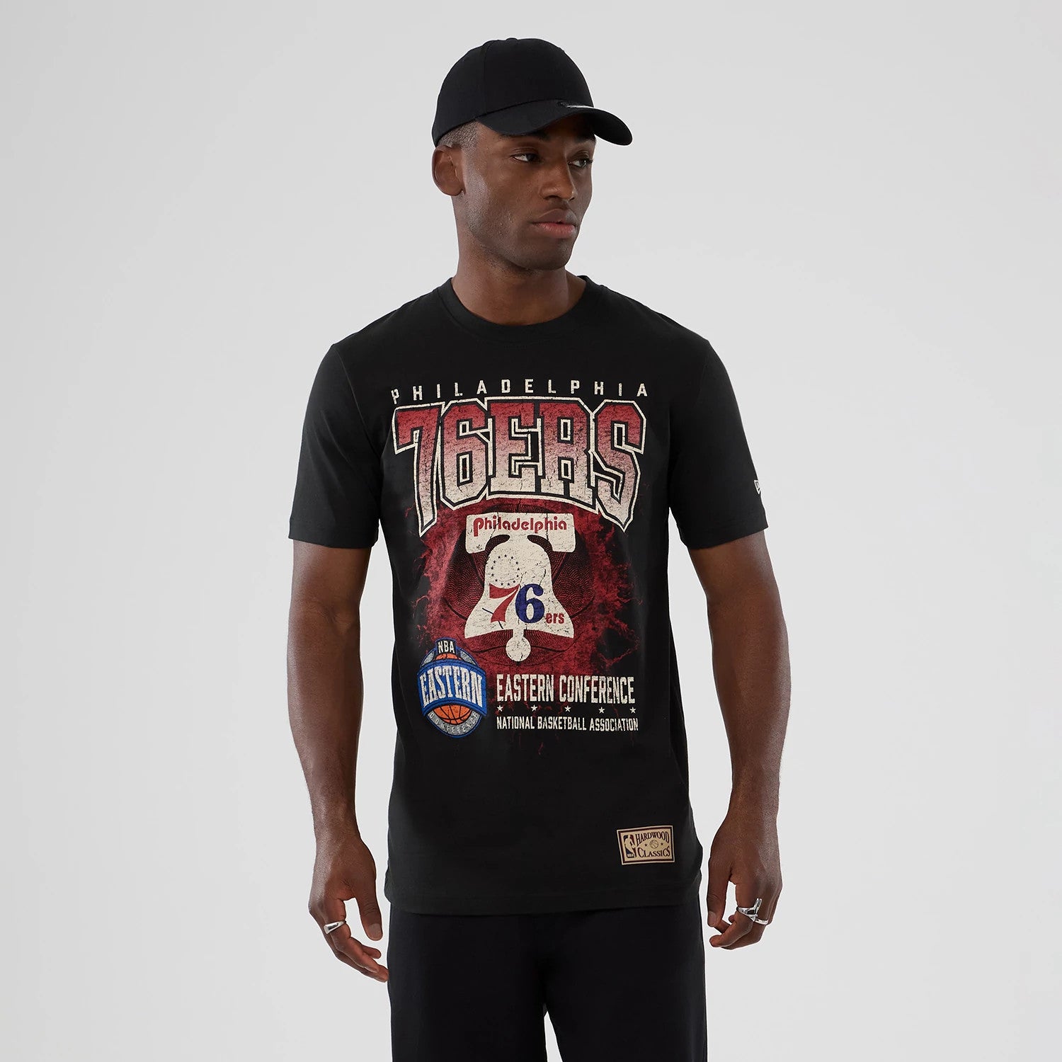 New Era Philadelphia 76ers NBA Hardwood Classic Black T-Shirt, front view on-model featuring a large printed 76ers graphic on the chest with Eastern Conference text and a woven Hardwood Classics patch near the hem.